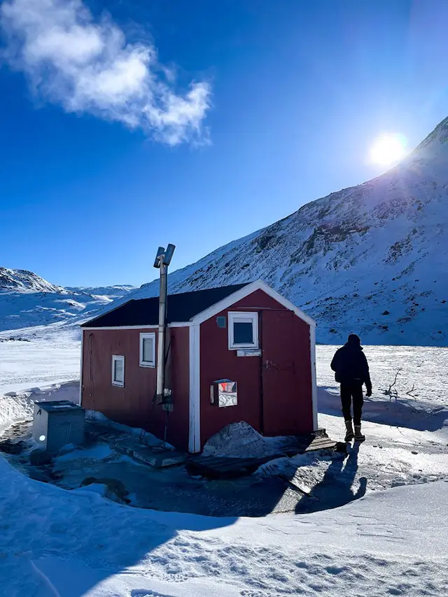 Červená nouzová chatka na trase Arctic Circle Trail v Grónsku, zalitá nízkým zimním sluncem. V okolí leží hluboký sníh a v dálce jsou vidět hory.