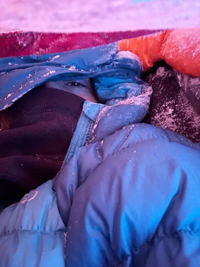 Detailní záběr na turistku zachumlanou v teplém spacáku a péřové bundě, které jsou pokryté jinovatkou během mrazivého rána na Arctic Circle Trail.