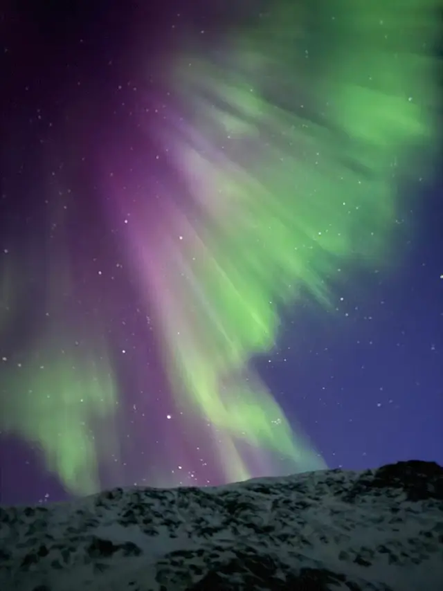 Zelená a fialová polární záře (aurora borealis) tančící na noční obloze plné hvězd nad zasněženou krajinou na trase Arctic Circle Trail v Grónsku.