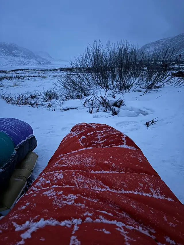 Pohled z oranžového zimního spacáku Rab Andes Infinium 800 pokrytého jinovatkou na zasněženou krajinu během přechodu Arctic Circle Trail v Grónsku