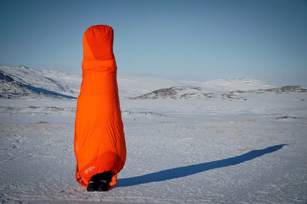 Testování zimního spacáku Rab Andes Infinium 800 v arktických podmínkách na Arctic Circle Trail. Oranžový spacák stojí na sněhu a demonstruje svůj objem a tepelnou izolaci.