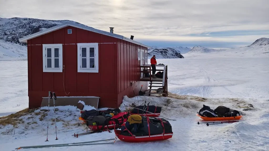 Červená nouzová chatka na trase Arctic Circle Trail v Grónsku, zalitá nízkým zimním sluncem. V okolí leží hluboký sníh a v dálce jsou vidět hory.